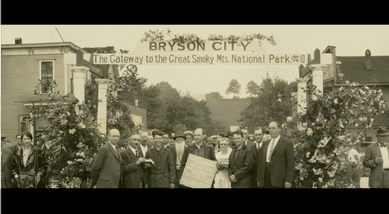 Screenshot from “A Life Reimagined: The George Masa Story” An old image of people standing in front of a banner that reads “Bryson City: The Gateway to the Great Smoky Mts. National Park”